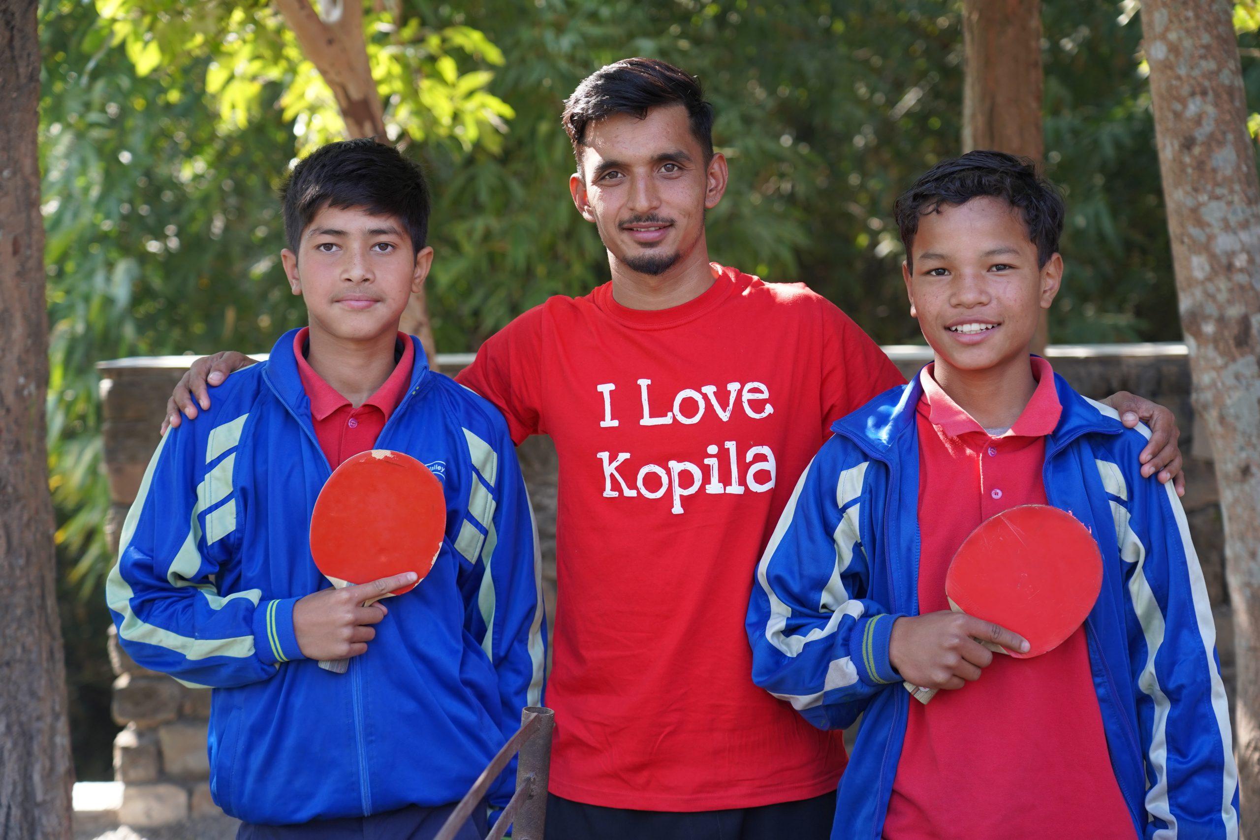 A man in a red "I Love Kopila" shirt stands outdoors with two boys who are holding table tennis paddles. The boys are wearing blue tracksuits with red shirts. Trees are visible in the background.