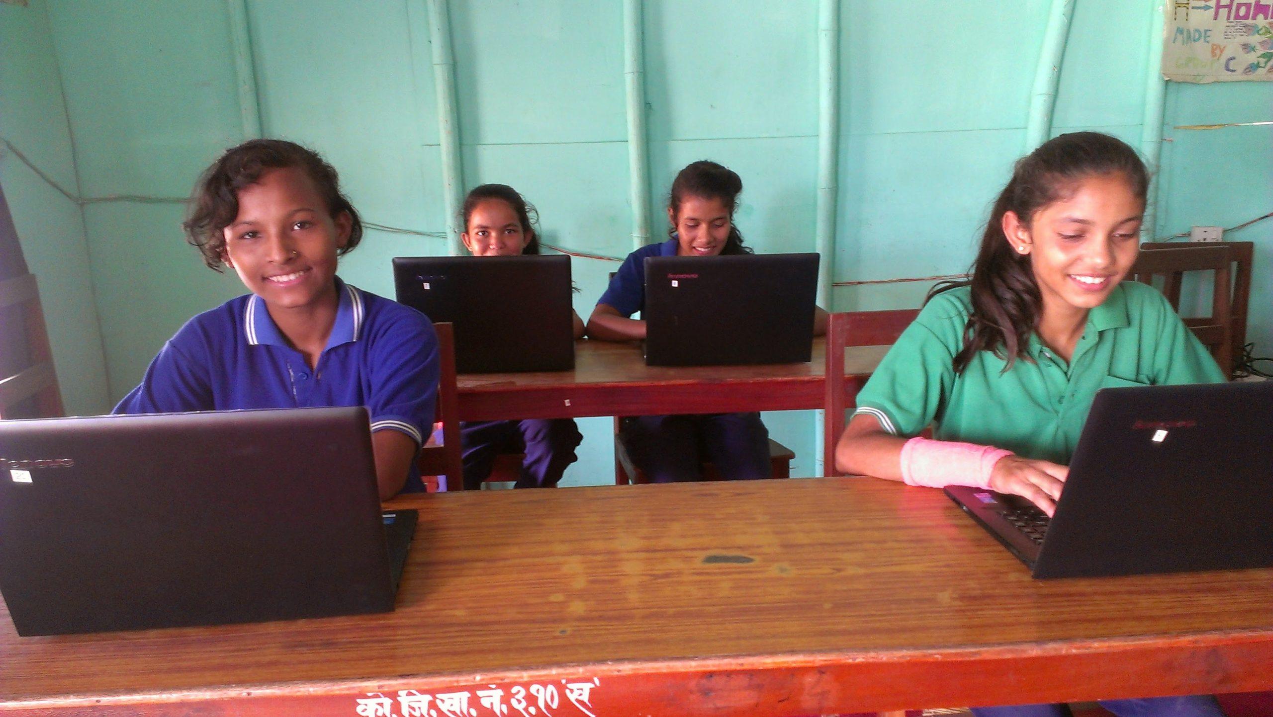 Four students sitting at desks in a classroom, each using a laptop. They are smiling and appear engaged in their work. The room has green walls, and the desks are wooden.