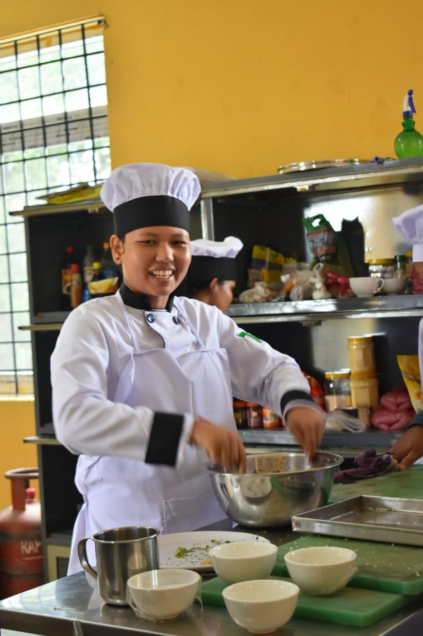 A smiling chef in a white chef's outfit is mixing ingredients in a kitchen. They are standing in front of a metal shelf filled with various cooking items, with bowls and utensils on the countertop.