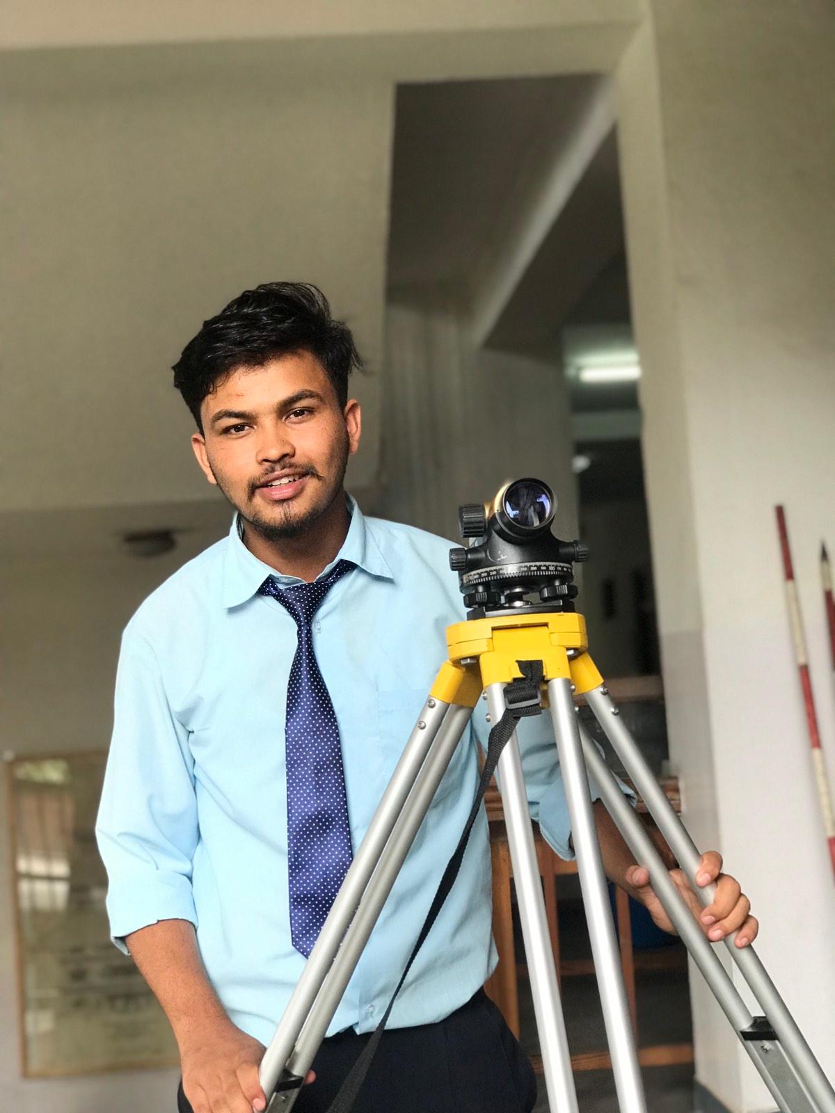 A man in a light blue shirt and tie stands indoors next to a tripod with a mounted surveying instrument, smiling at the camera.