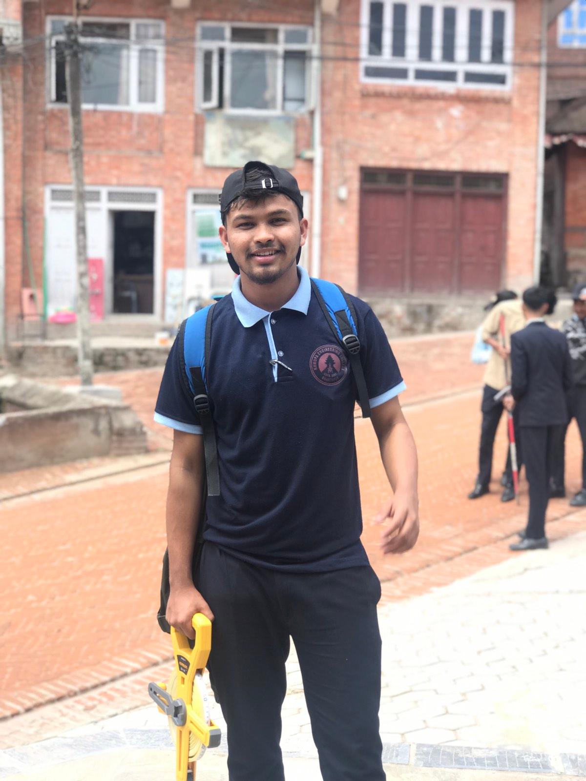 A person wearing a navy blue polo shirt and black cap stands outdoors, smiling. They hold a yellow tool and have a backpack. In the background, there are brick buildings and other people walking.
