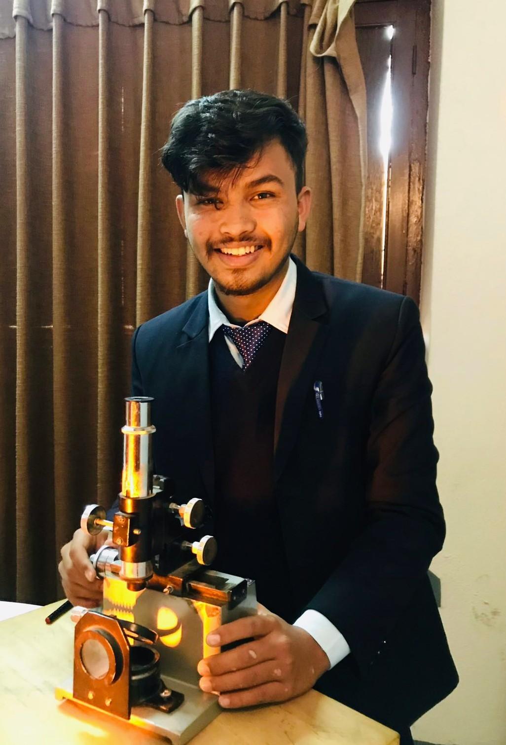 A person smiling and standing behind a microscope on a table, wearing a suit and tie. The background features a brown curtain.