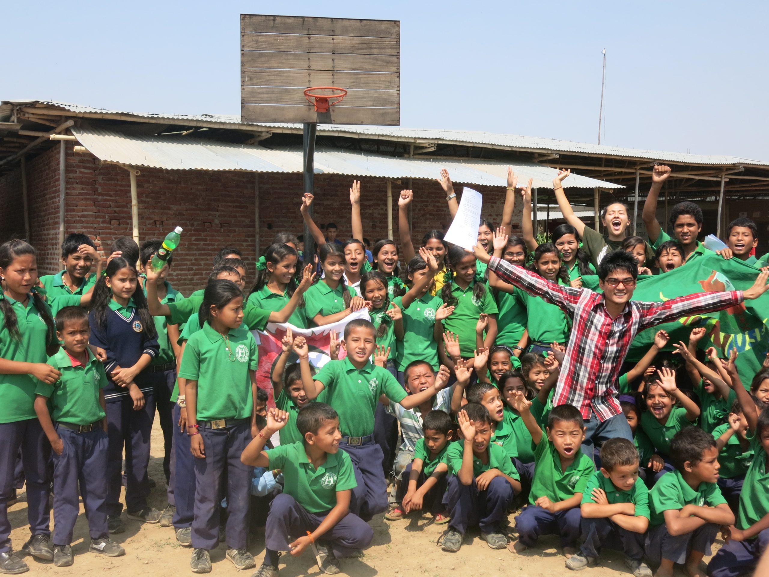 A large group of children wearing green uniforms cheer enthusiastically, raising their arms in celebration. An adult in a plaid shirt stands in front, stretching out their arms with a big smile. They are outside, with a building in the background.