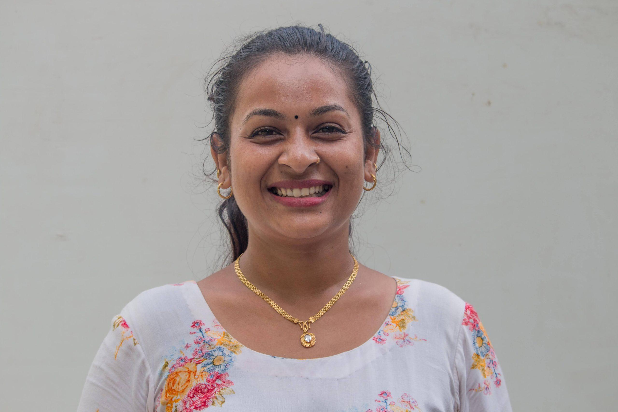 A woman with a bright smile, wearing a floral white top and gold jewelry, stands in front of a plain light gray background. Her hair is tied back, and she has a small bindi on her forehead.