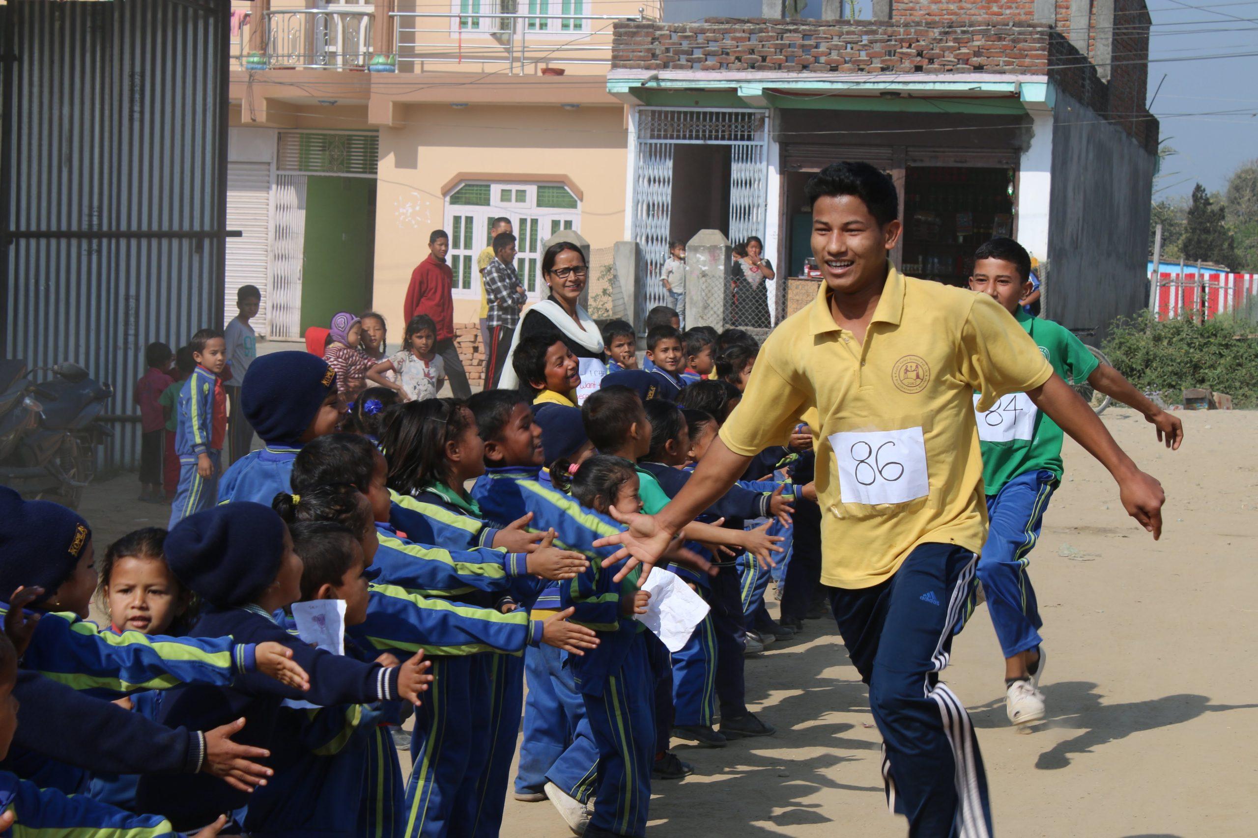 A man in a yellow shirt, wearing a race number, runs past cheering children in blue uniforms. The scene takes place on a sunny day, with a row of houses in the background and onlookers watching.