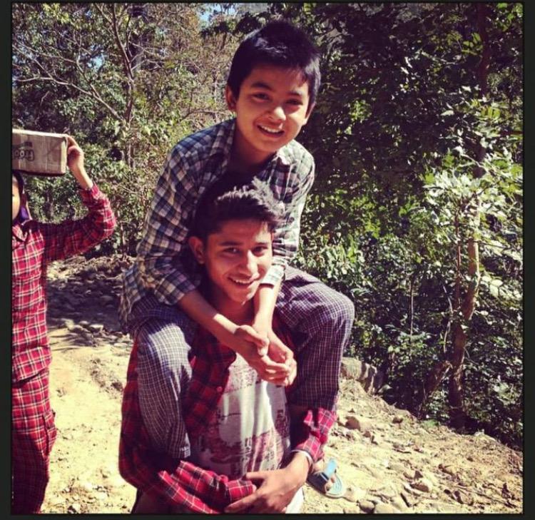 A boy carrying another boy on his shoulders, both smiling and dressed in casual clothes. They are outdoors on a sunny day, surrounded by greenery. Another child in the background carries a container on their head.