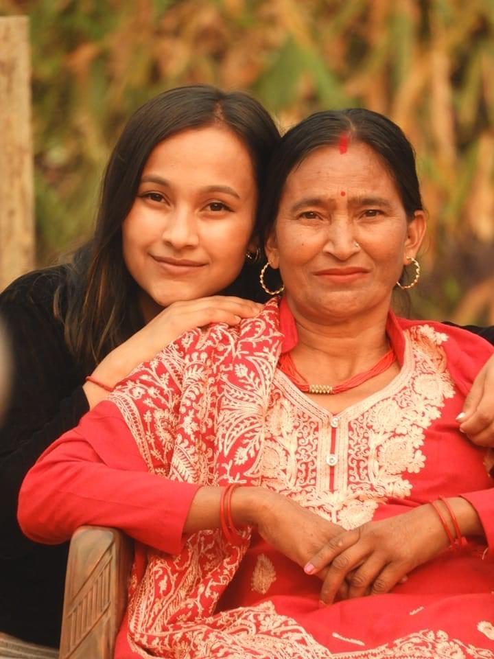 A young woman and an older woman sit closely together outdoors, smiling at the camera. The older woman wears a red and white traditional outfit. They appear happy and relaxed.
