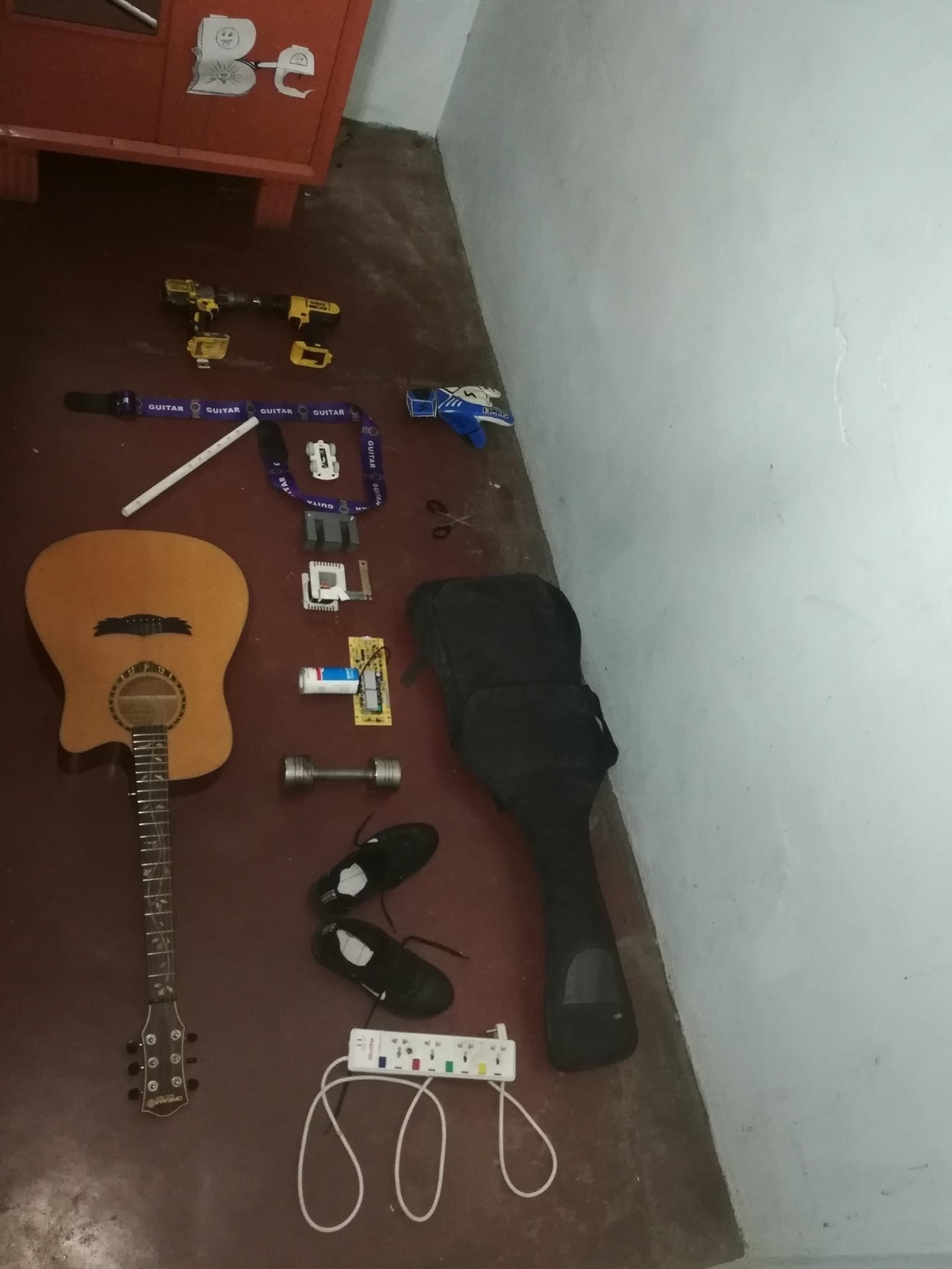 An acoustic guitar, a guitar case, a pair of black shoes, a power strip, a bike lock, drill tools, weights, paint roller, and a box of screws are laid out on a brown floor next to a red cabinet against a pale wall.