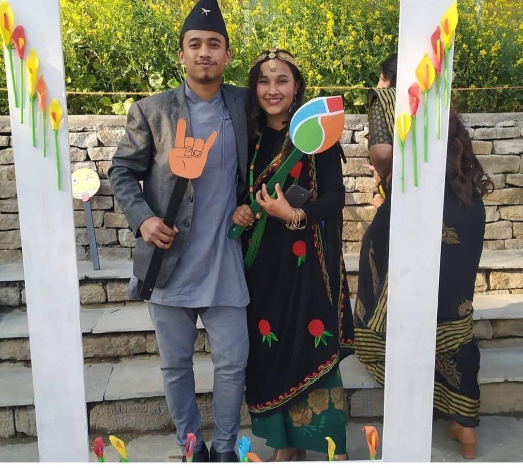 A man and woman dressed in traditional attire stand together, holding paper cutouts with symbols. They are framed by a decorated archway, with a background of yellow flowers and stone steps. The mood is festive and celebratory.