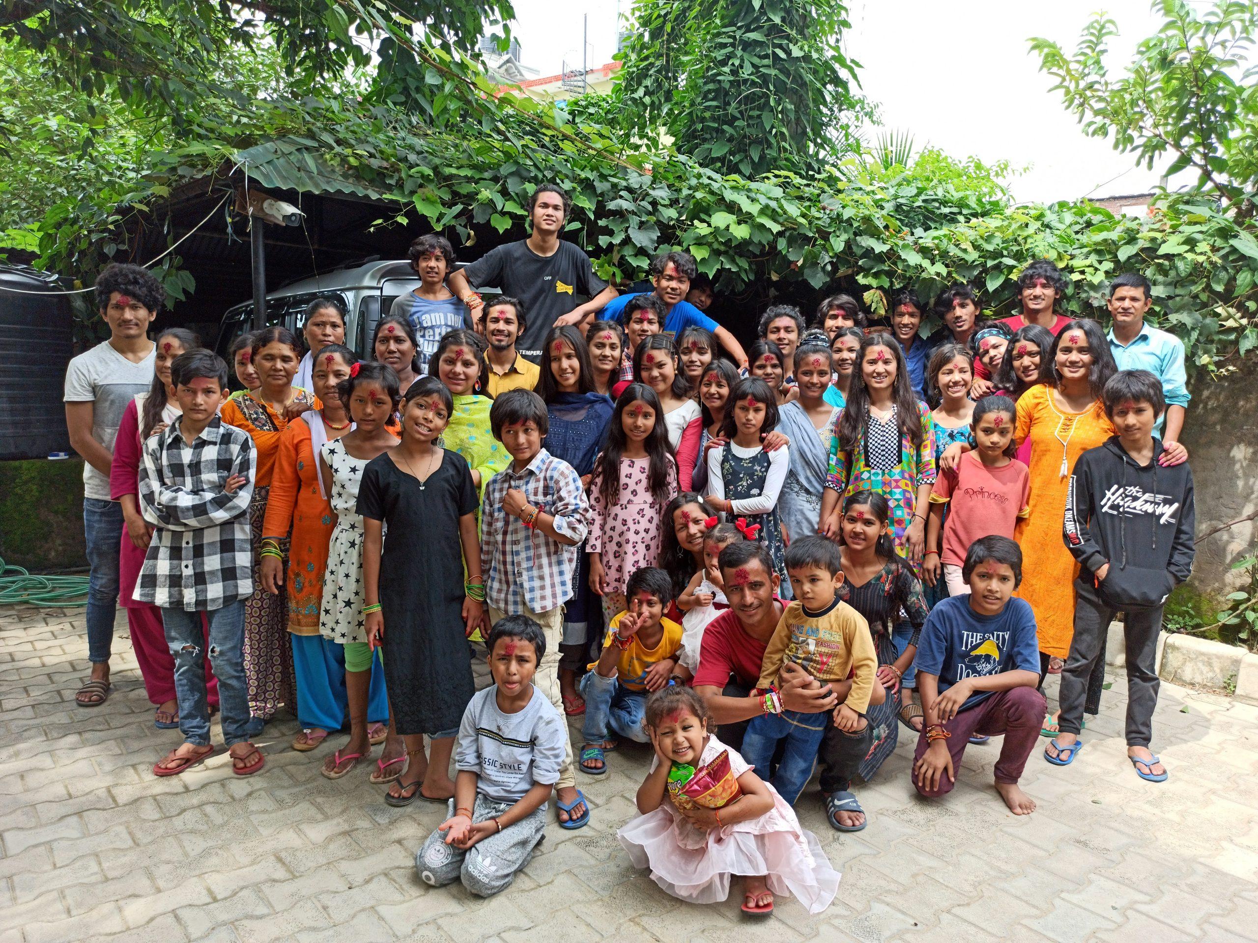 A large group of smiling children and adults posing together outdoors. The background features greenery and some parked vehicles. The diversity of clothing adds color to the lively scene.