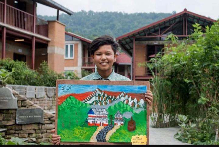 A person stands outdoors, smiling and holding a colorful painting of a mountainous landscape with a house and trees. Behind them are brick buildings and greenery under a cloudy sky.