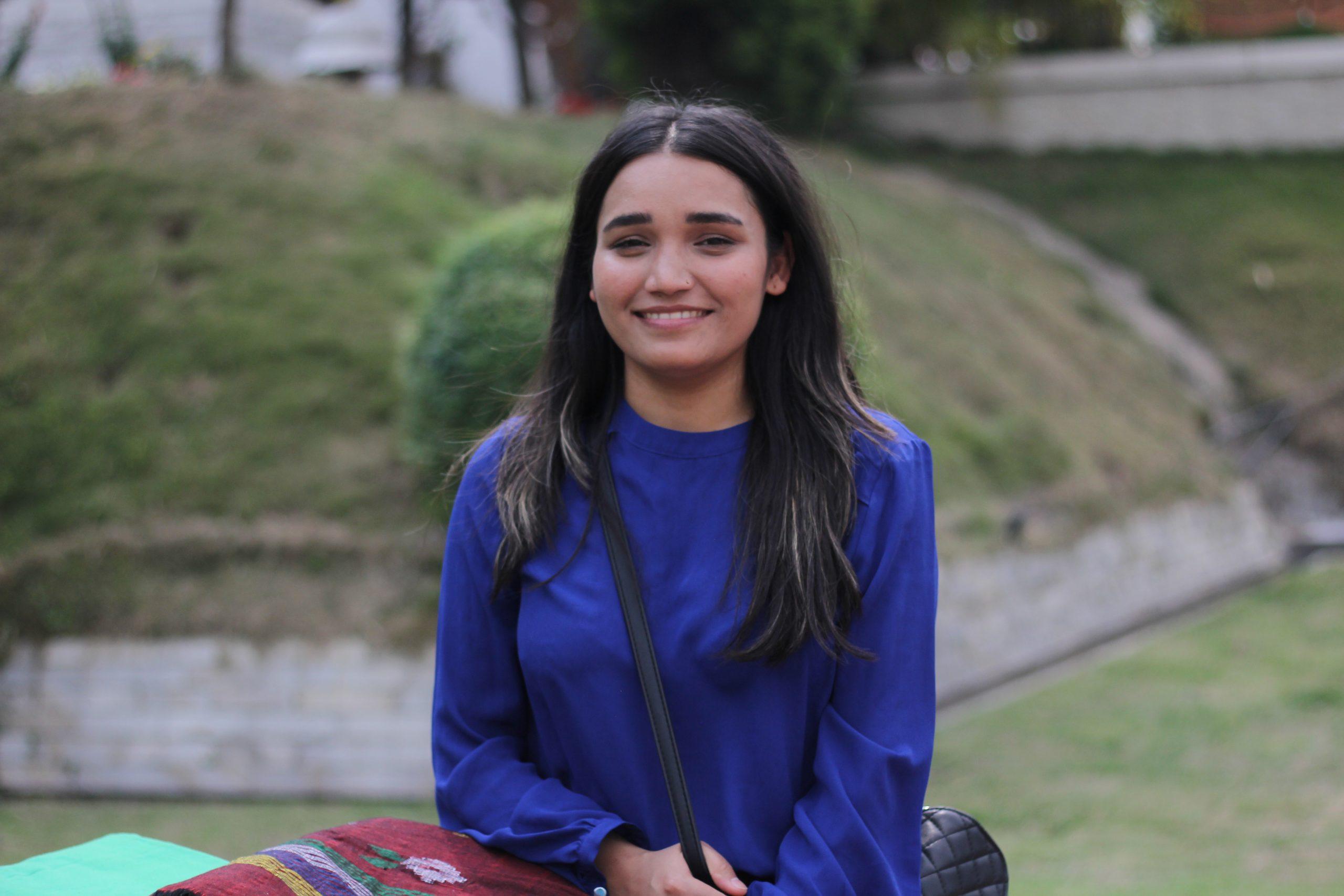 A woman with long dark hair in a blue shirt smiles while sitting outside. She has a black shoulder bag, and a grassy hill is visible in the blurred background.
