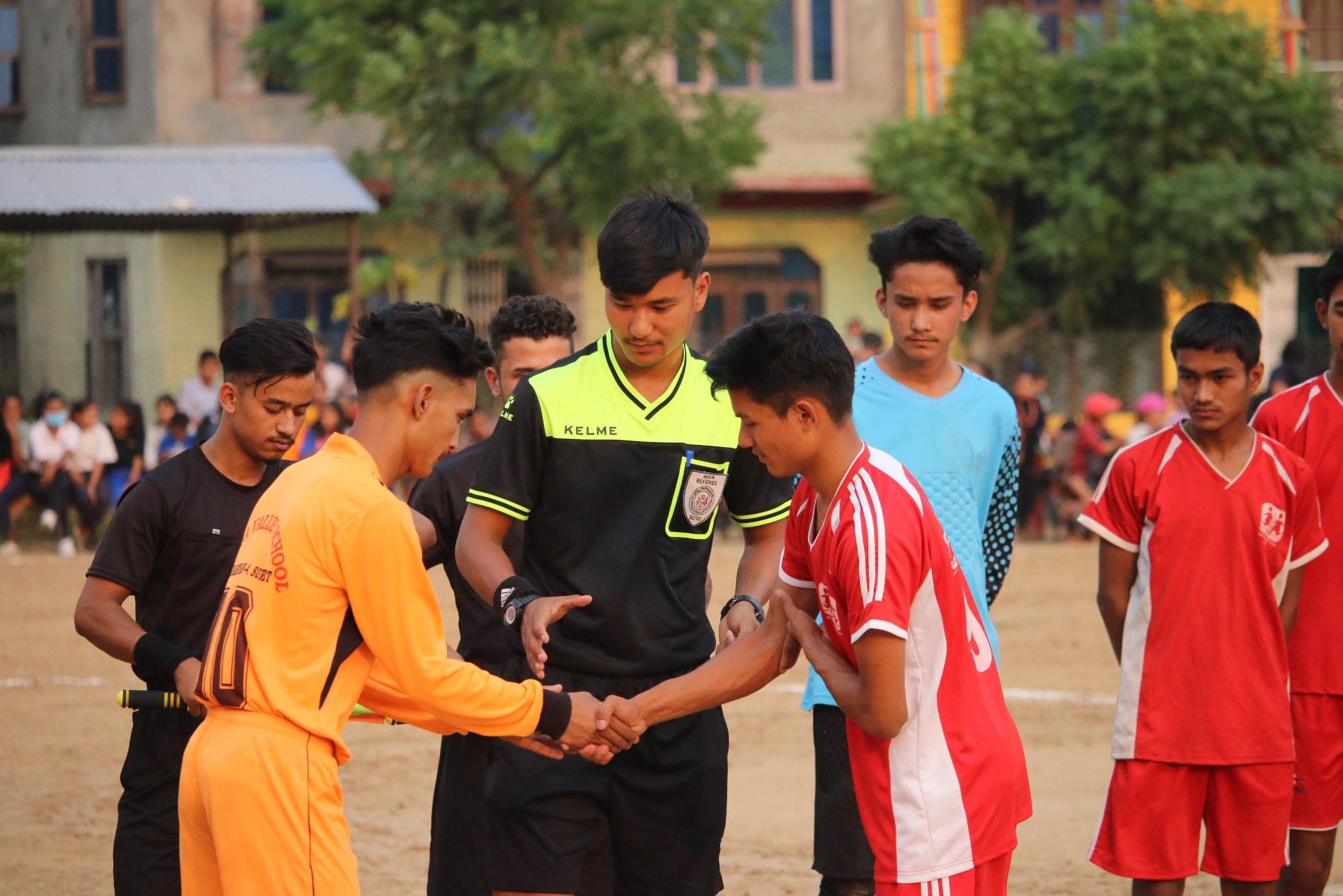 A referee stands on a soccer field with players from two teams, one in orange and the other in red, shaking hands before a community match. Other players and spectators are visible in the background.