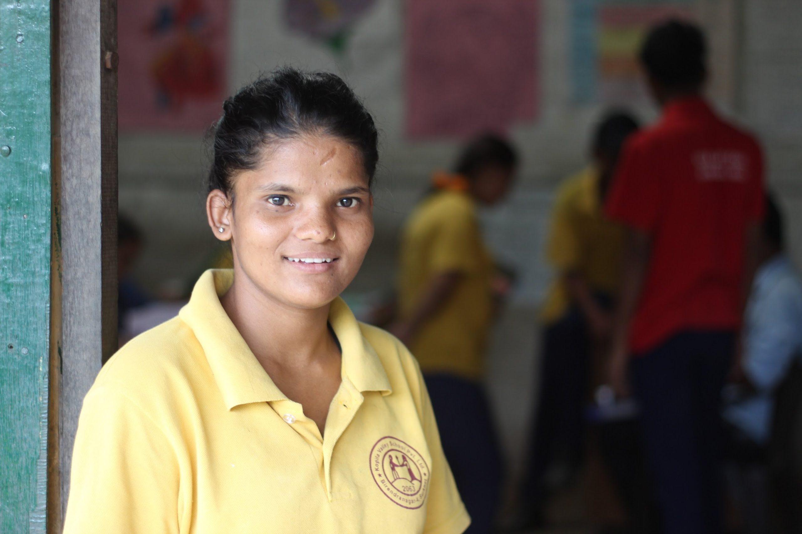A woman in a yellow shirt stands indoors, smiling at the camera. In the background, several people dressed in yellow and red shirts are engaged in various activities, with colorful posters on the walls.