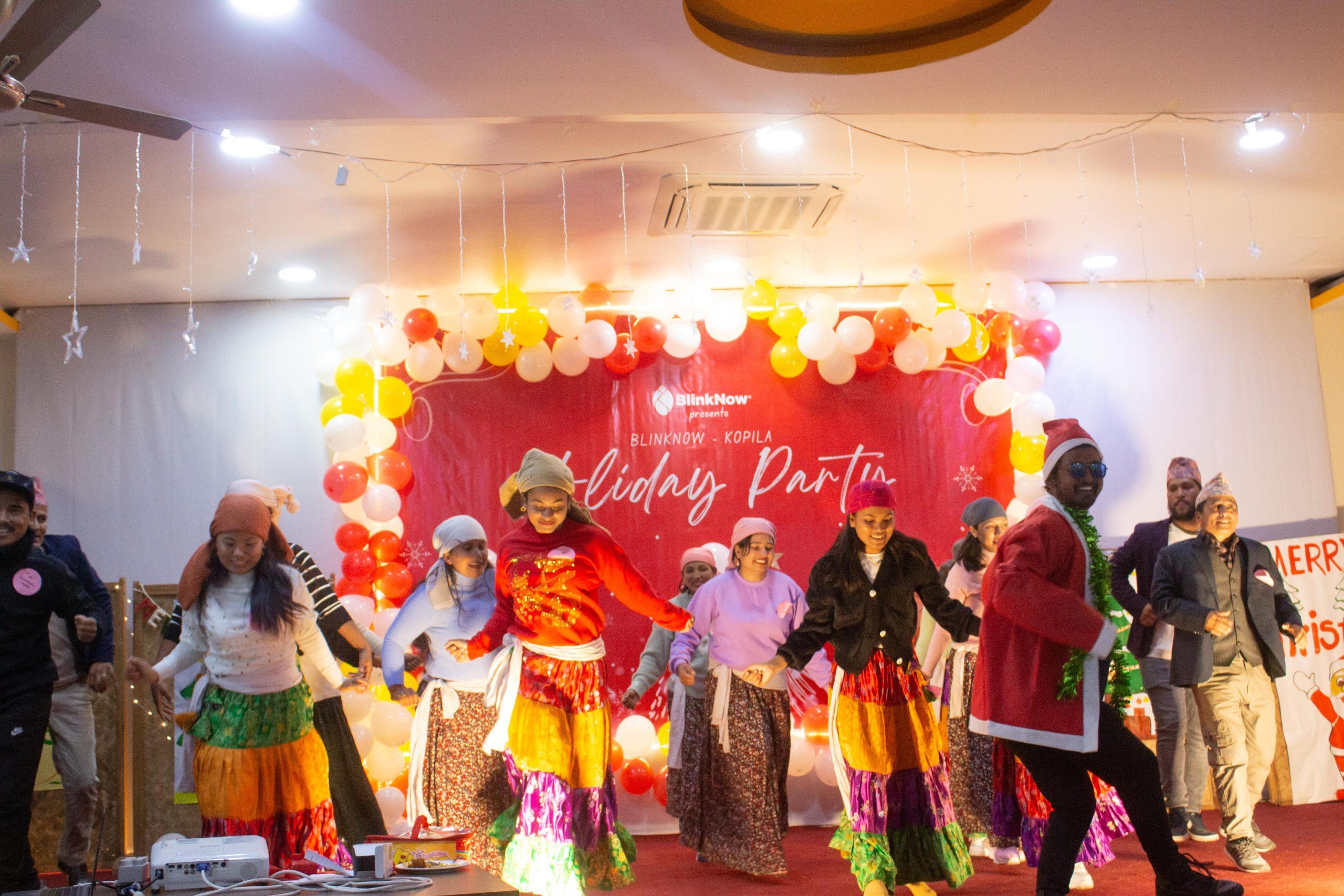 A lively group of people dances on a decorated stage at a holiday party. They are wearing colorful skirts and hats. The background features festive balloons and a red banner with the text "Holiday Party.