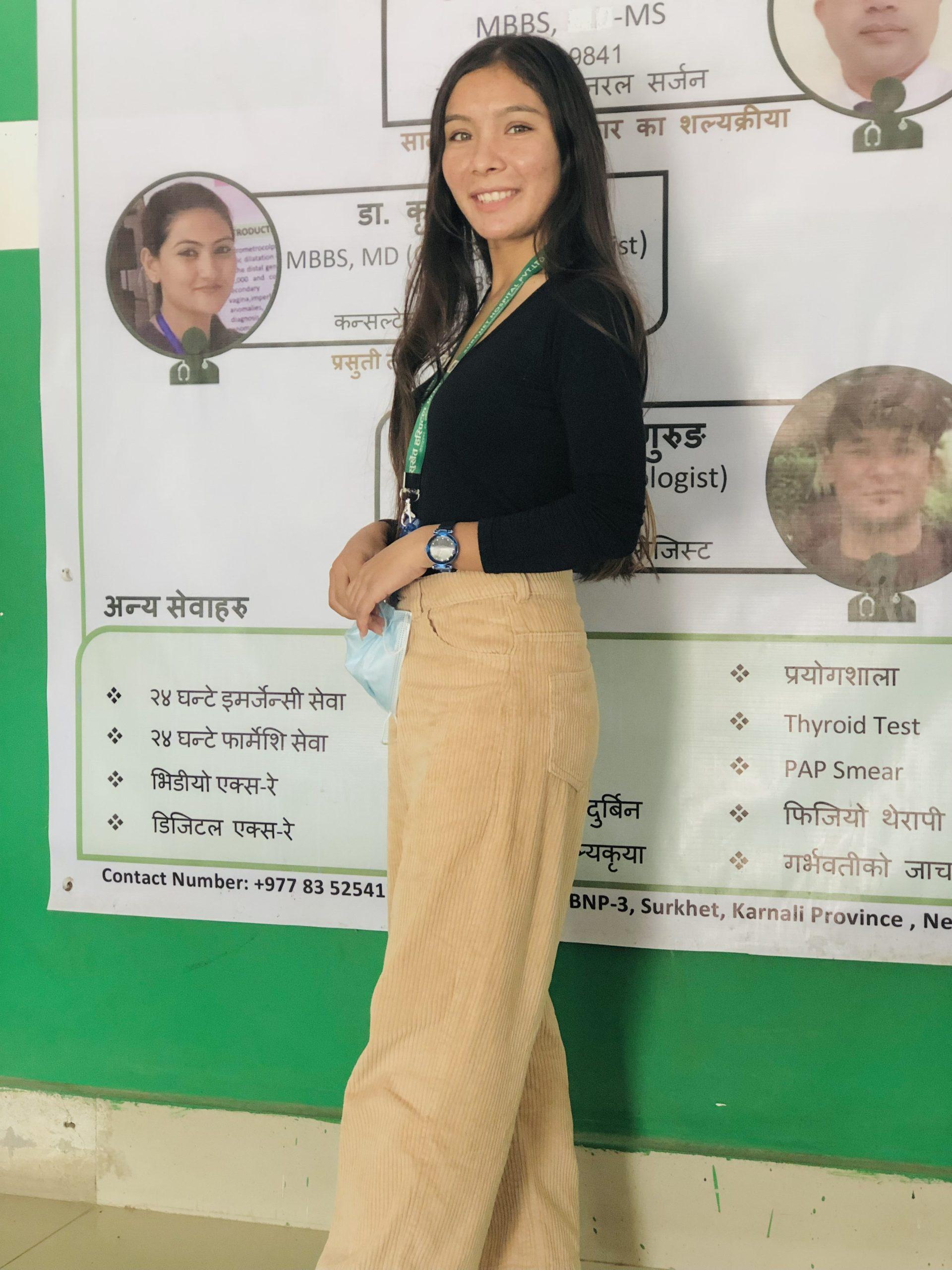 A woman stands in front of a poster with medical information. She is wearing a black top, beige pants, a lanyard, and a watch. The background shows various text and photographs of medical professionals.