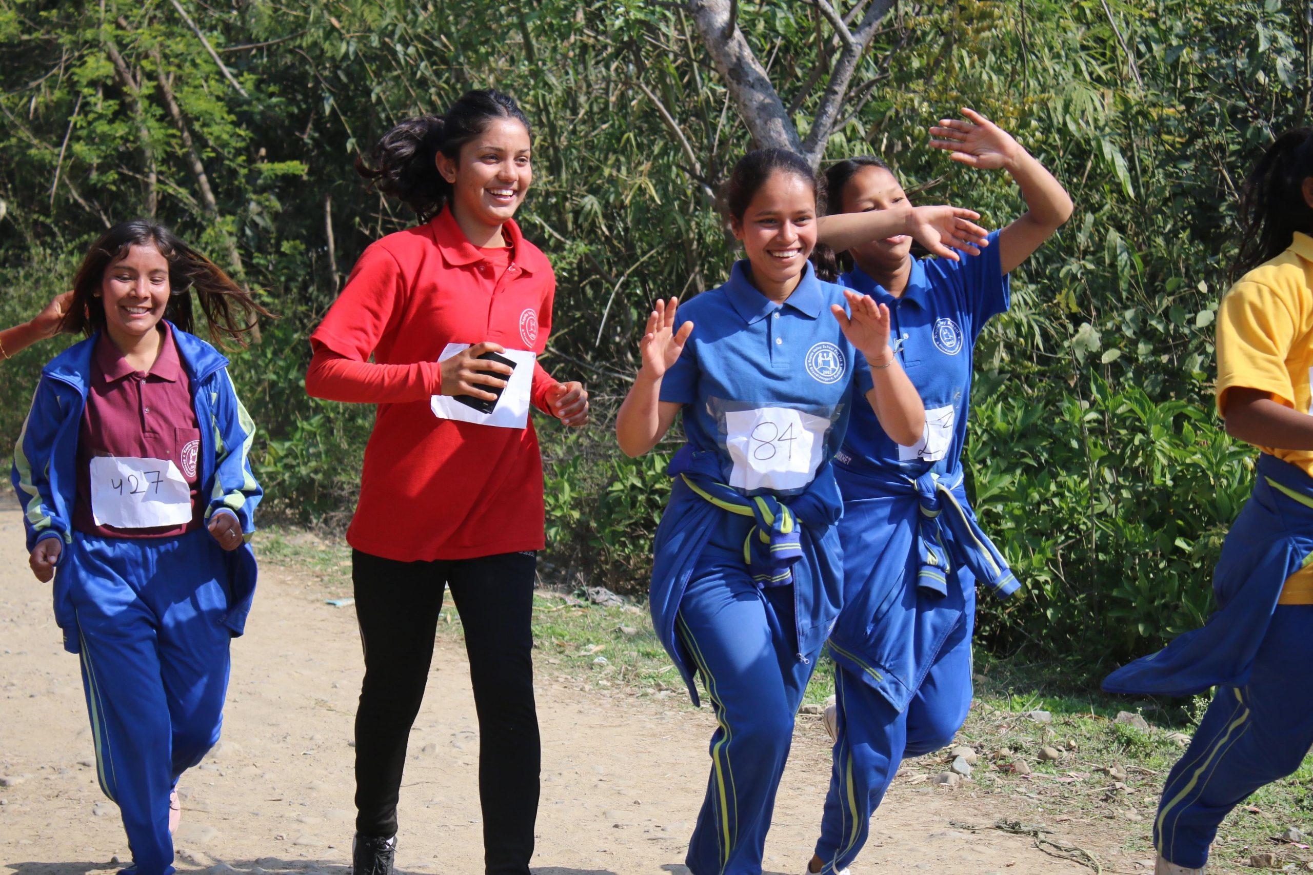 A group of young people in athletic clothing are jogging on a dirt path. They are wearing numbered bibs and appear cheerful. Tall trees are visible in the background, and the day is sunny.
