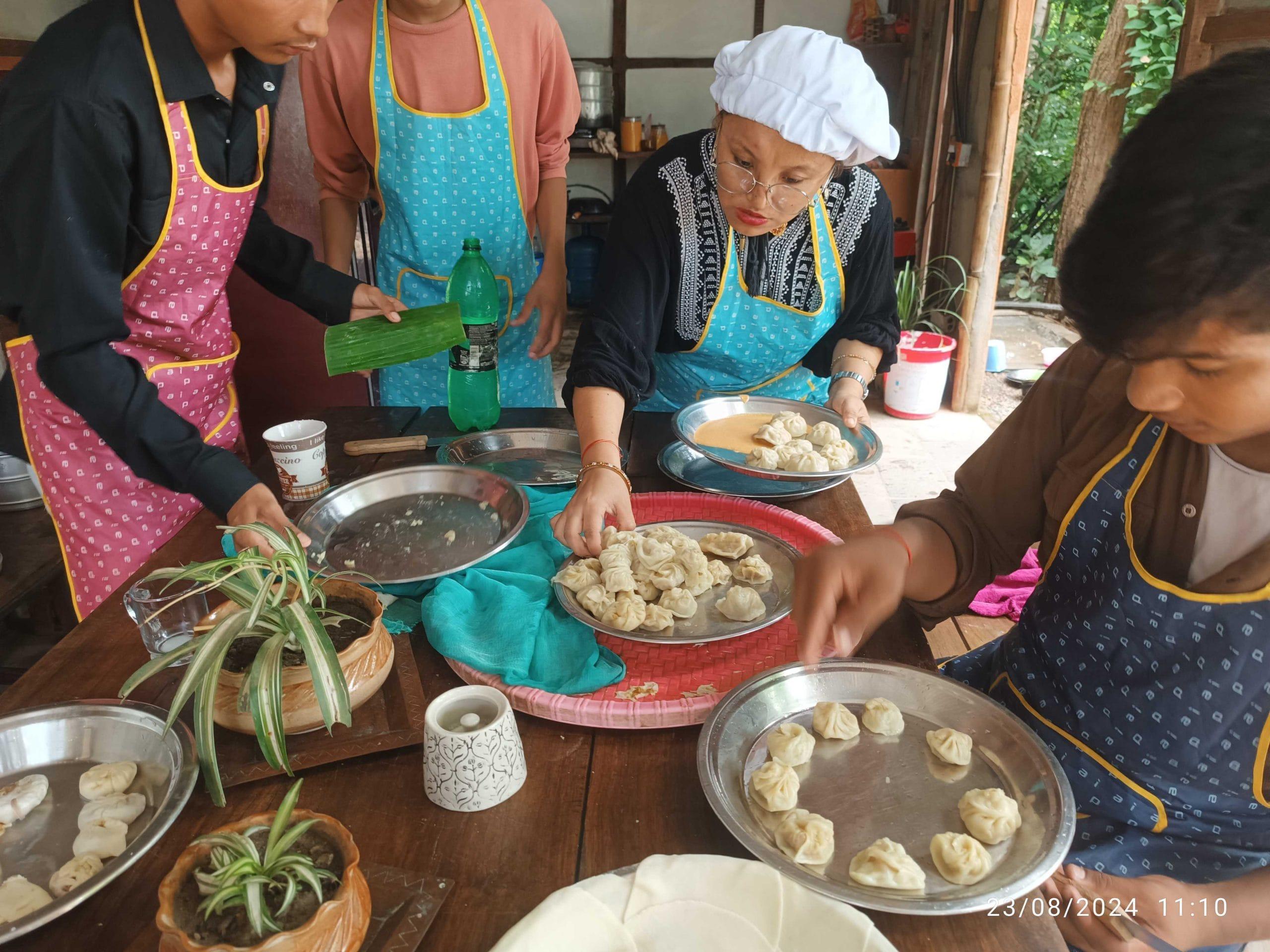 A group of people in aprons and a chef's hat are making dumplings around a wooden table. Various kitchen items and plants are on the table. The setting appears to be a cozy kitchen or cooking class.
