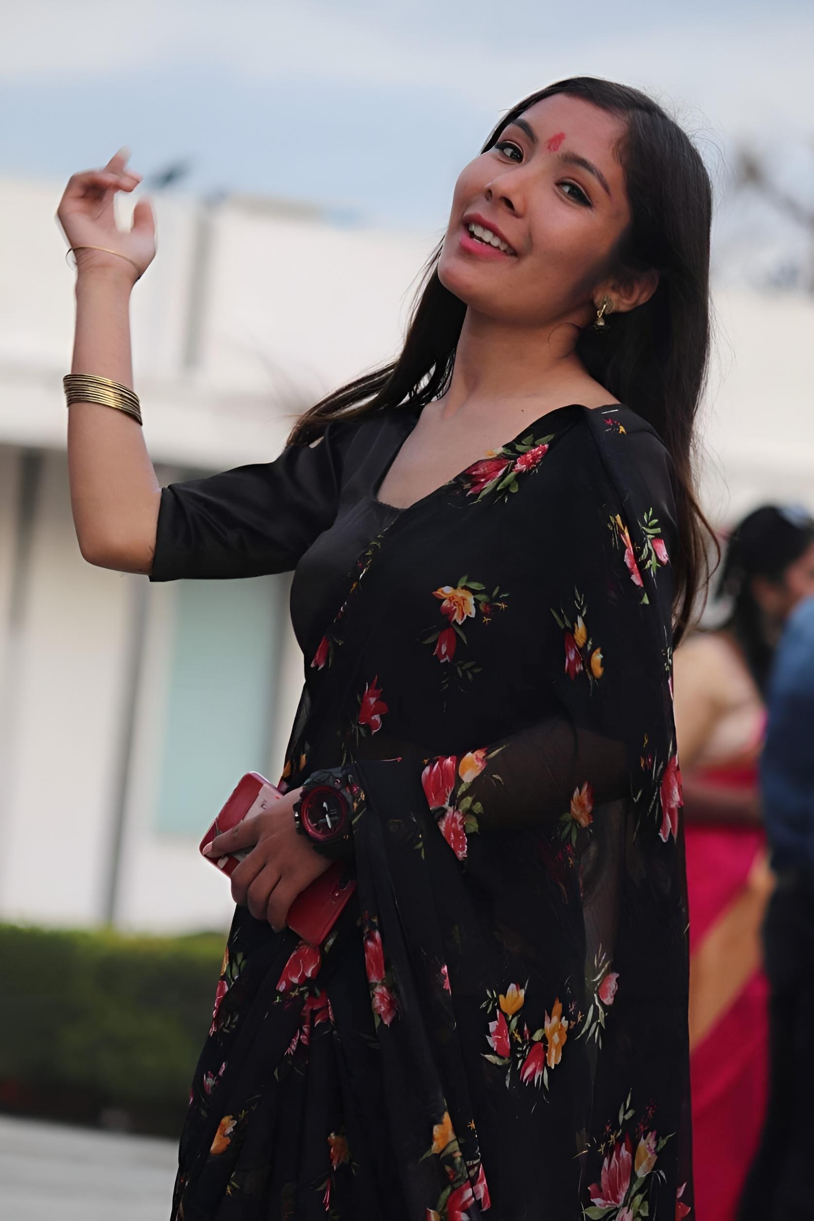A woman wearing a black saree with floral patterns and a red bindi smiles while holding a phone. She is outdoors, with a blurred background of greenery and a white building.