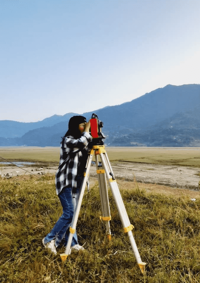 A person wearing a black hat and plaid shirt uses a theodolite on a tripod, surveying a scenic landscape with hills and open fields under a clear sky.