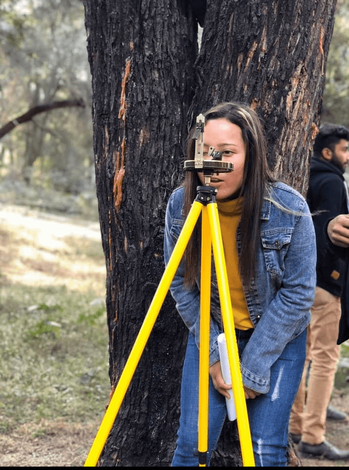 A person in a denim jacket is using a tripod-mounted theodolite for surveying, standing next to a tree in a wooded area. Another person is in the background.