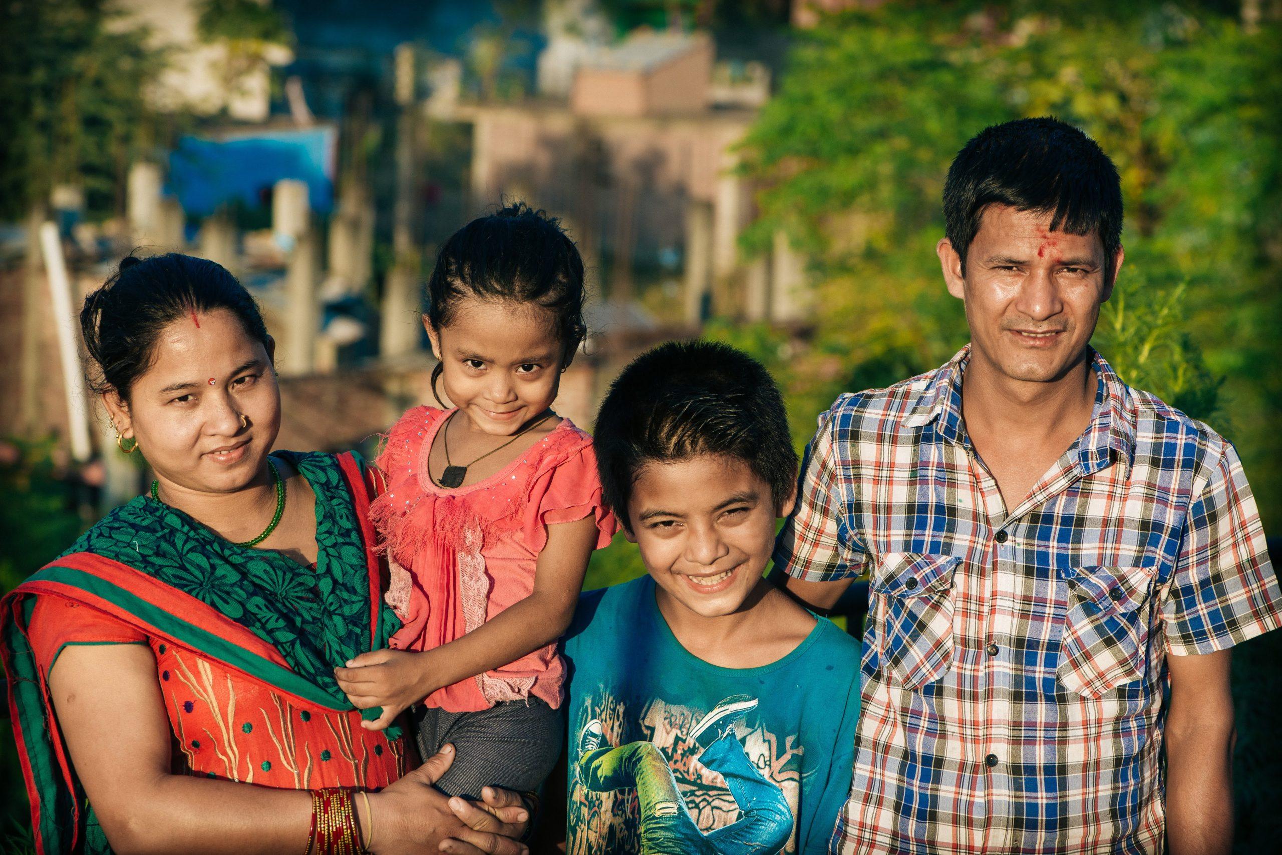 A family of four, including a mother, father, and two children, stands outdoors smiling. The mother is wearing a red and green saree, holding a young girl in pink, while the father in a checkered shirt stands next to a boy in a blue shirt.