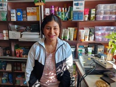A woman in a black and white jacket sits in a shop with shelves stocked with various cosmetic and skincare products. She is smiling, and there is a table with more products in front of her.