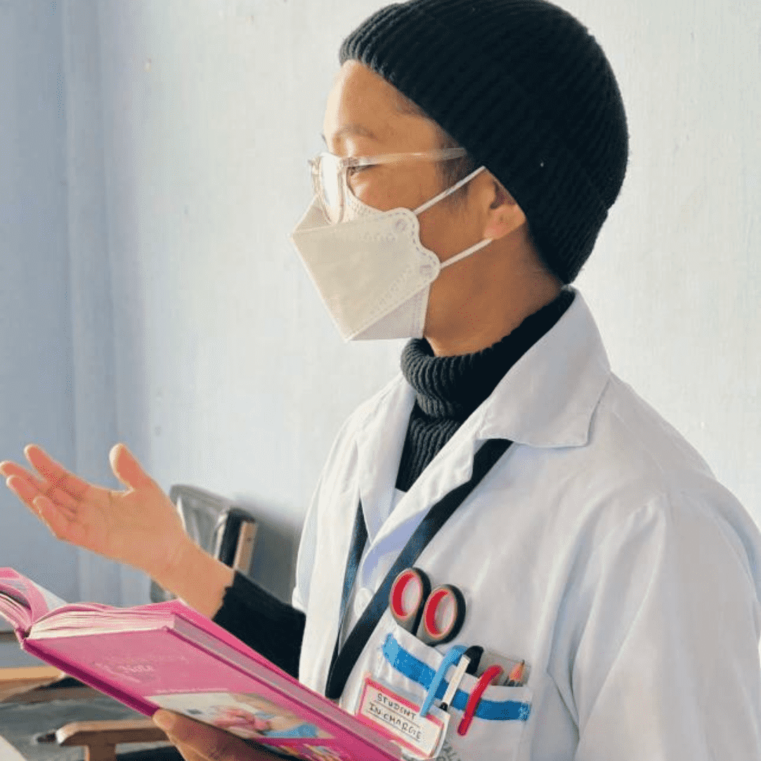 A person wearing a mask, a black beanie, and a white coat with pens in the chest pocket holds a pink notebook, gesturing with their other hand. They appear to be speaking in a classroom setting.