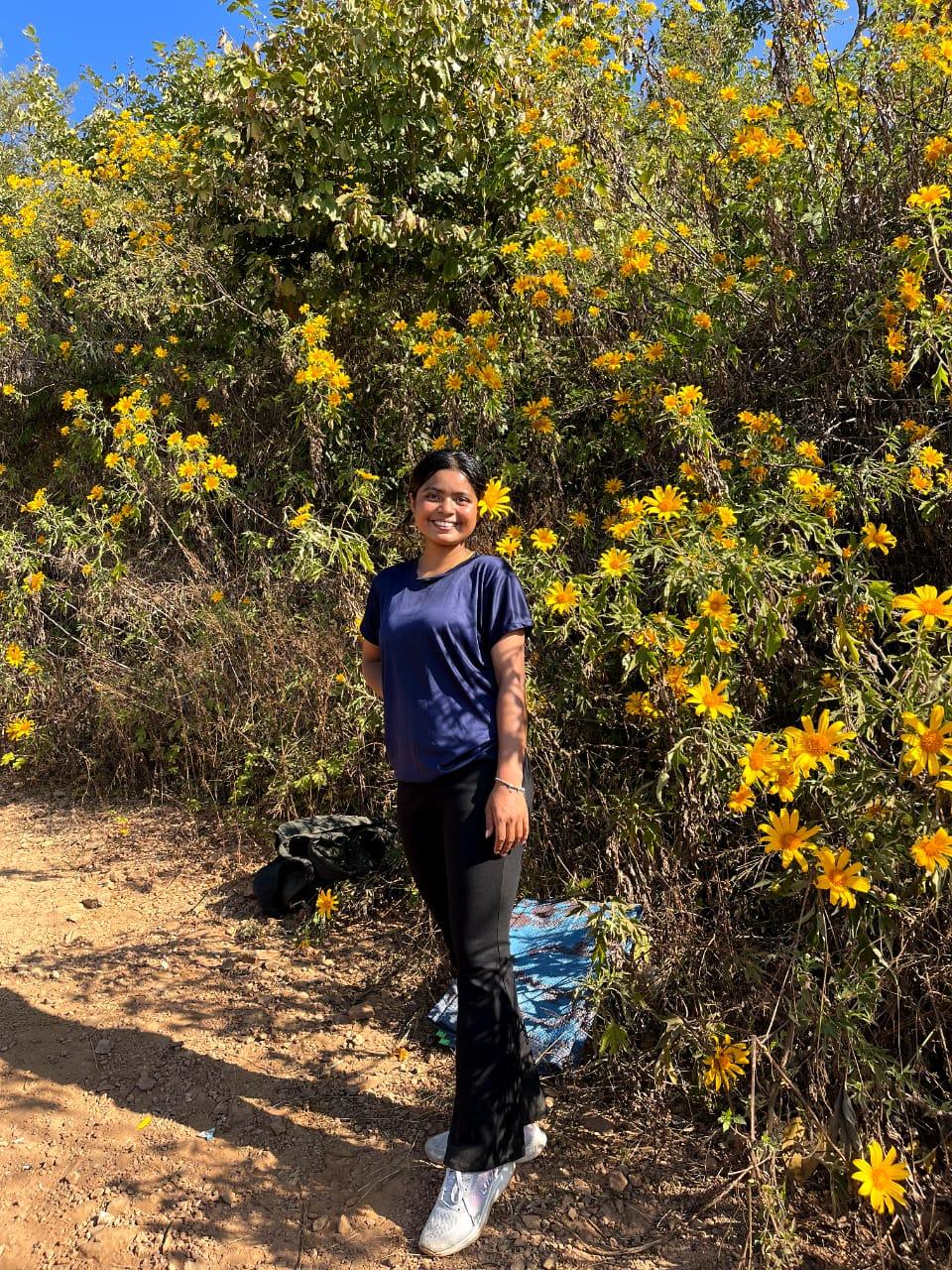 A person in a blue shirt and black pants stands smiling among vibrant yellow flowers on a sunny day. The ground is sandy, and lush greenery surrounds the scene.
