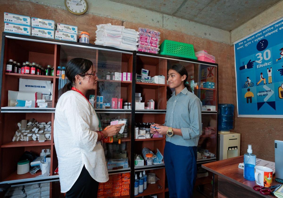 Two people are standing in a pharmacy. One is holding medication, and they are engaged in conversation. Shelves with various medical supplies and a poster on health tips are visible in the background.