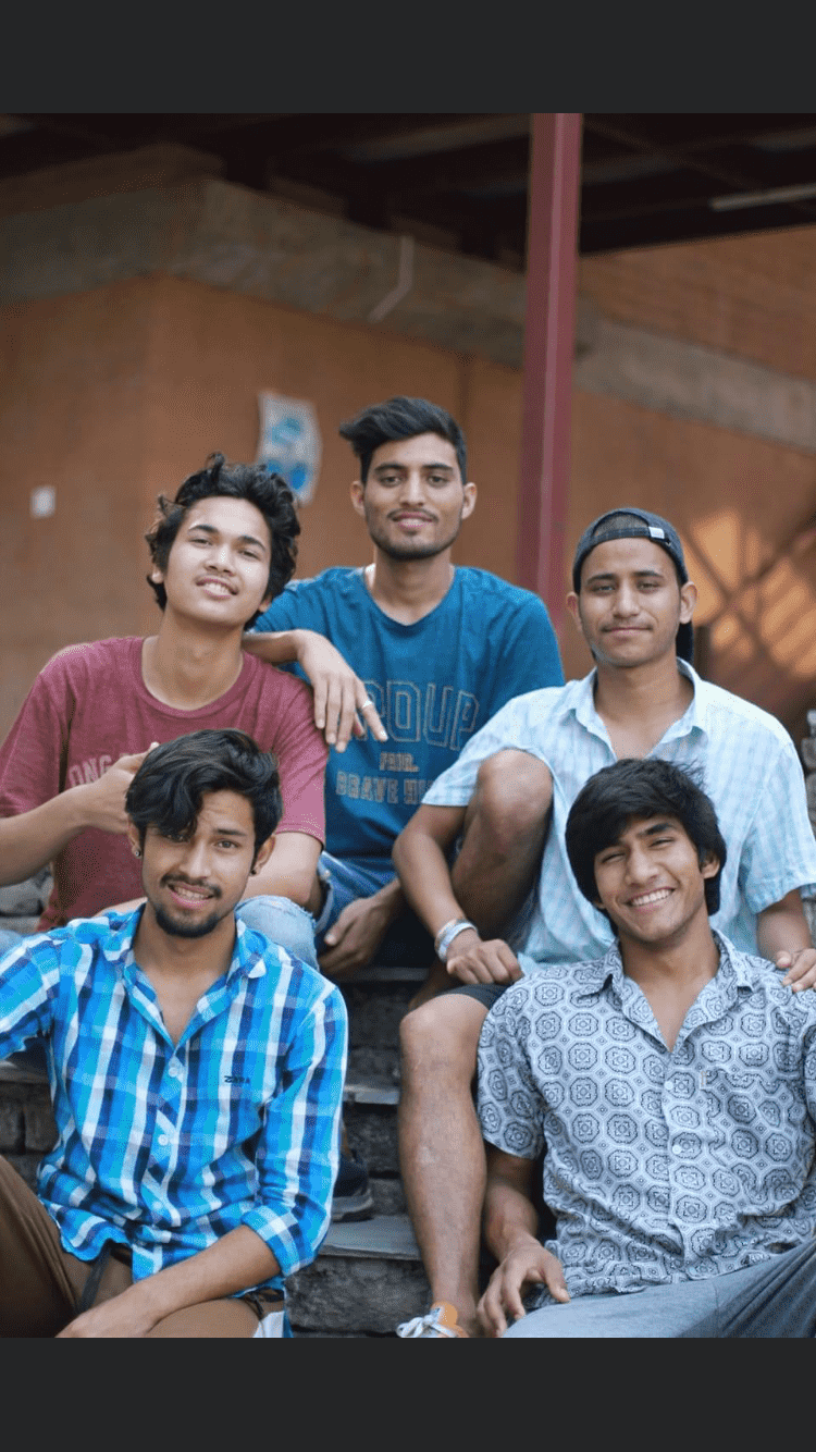 A group of five young men sitting on outdoor steps, smiling at the camera. They are casually dressed and appear relaxed. A brick building with a poster is in the background.