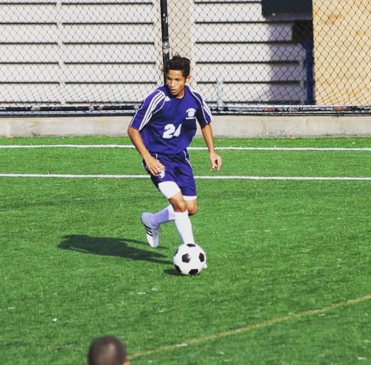 A young soccer player in a blue jersey with the number 21 controls the ball on a green field, while running. He's focused on the game, with a chain-link fence and building in the background.