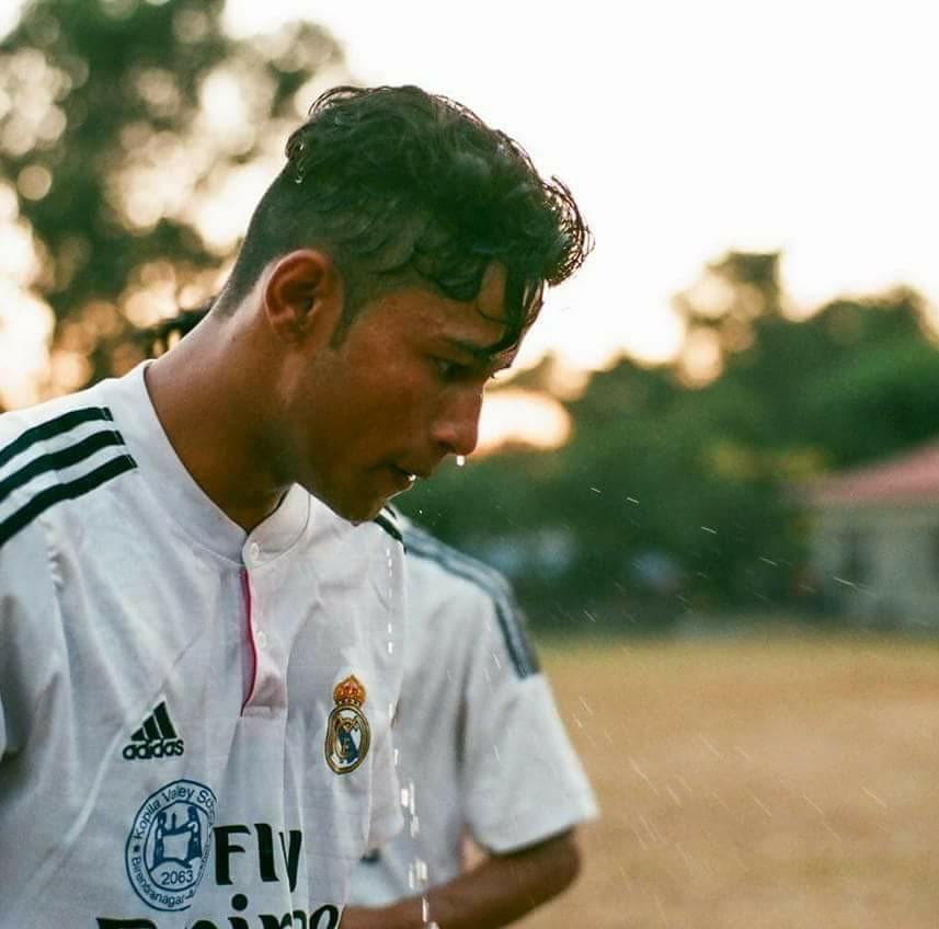 A soccer player in a white jersey with a crest, likely during practice, stands outside as water drips down his face. The background is a sunlit, blurred field with trees and a building.