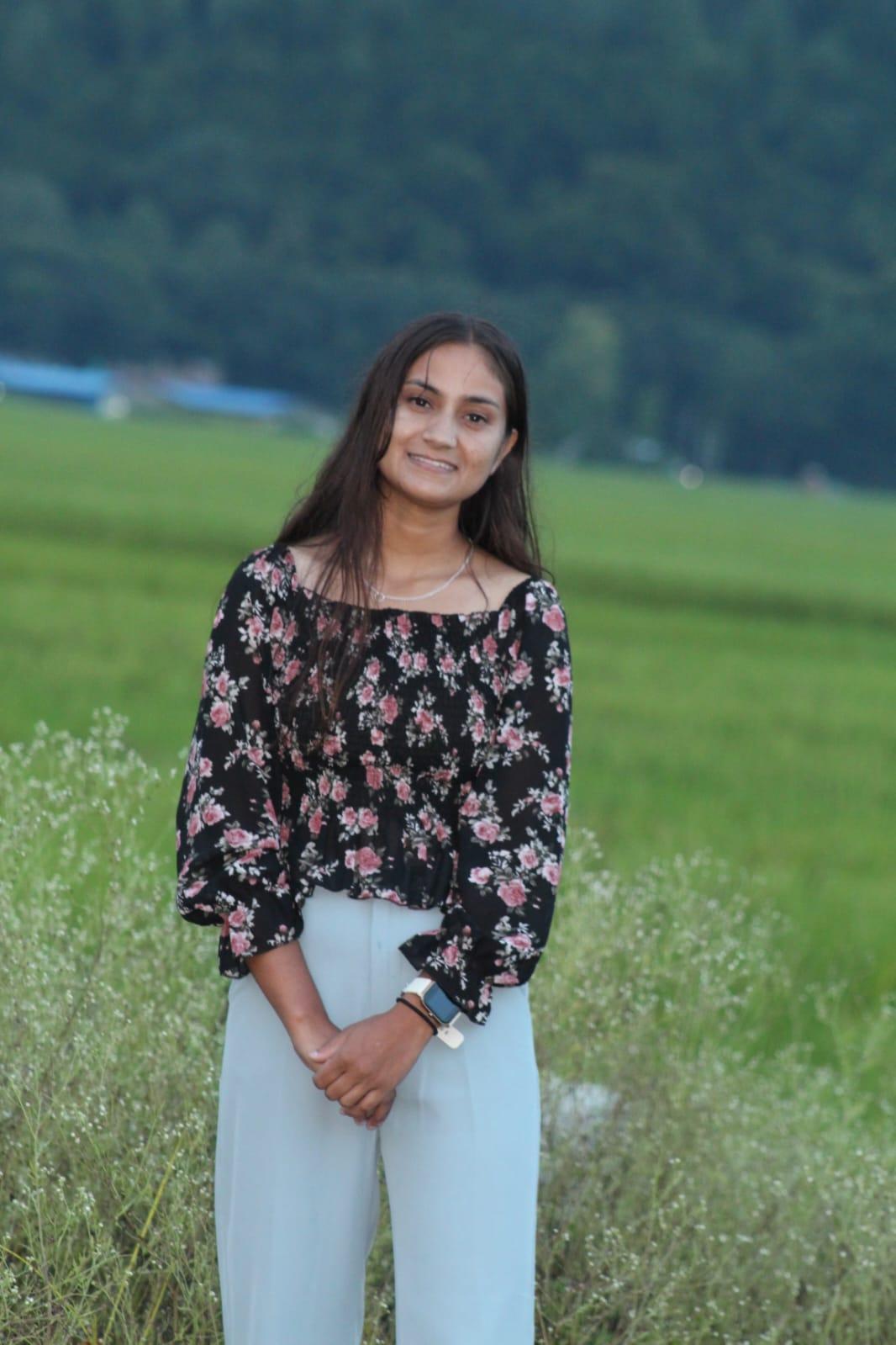 A person with long hair is standing in front of a lush green field, wearing a floral off-shoulder top and light-colored pants. A forested area is in the background.
