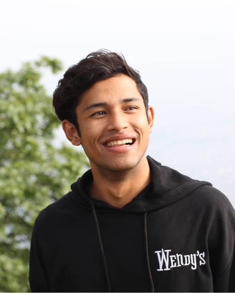 A person smiling, wearing a black hoodie with a "Wendy's" logo. The background features green foliage.