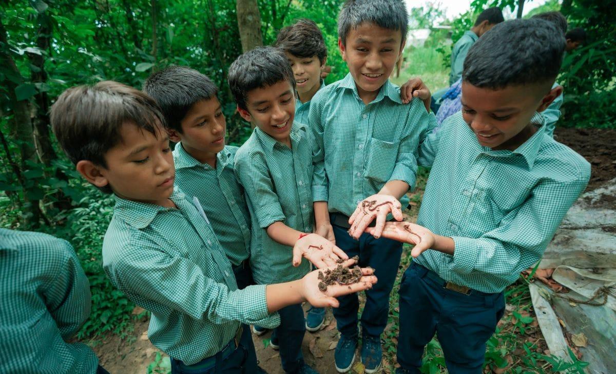 A group of boys in green uniforms eagerly examine earthworms in their hands while standing in a lush, green outdoor setting. They appear excited and curious as they explore nature together.