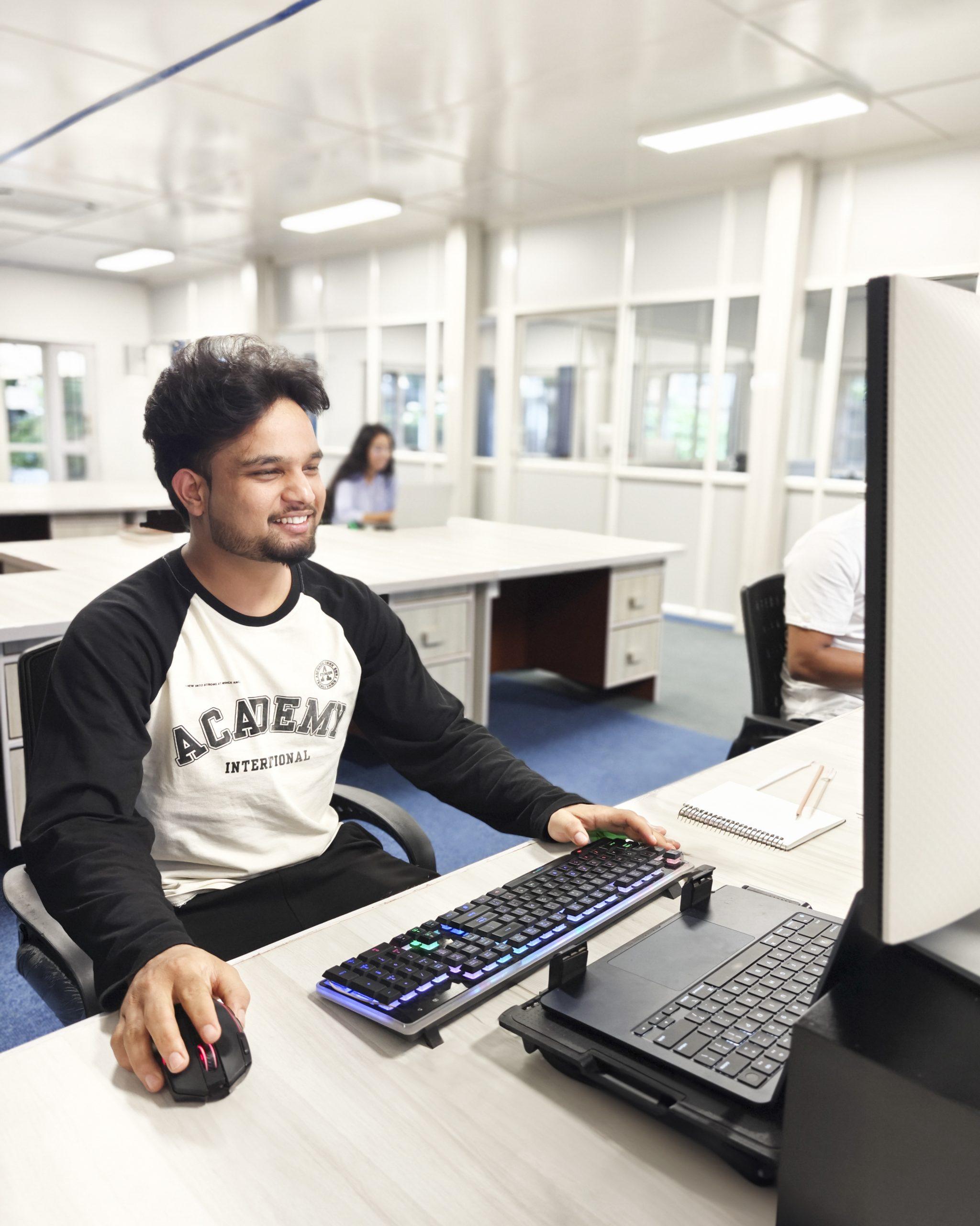 A person with dark hair and a beard is smiling while working on a computer in an office. Another person is seated at a desk in the background. The office has white desks and partitioned spaces.