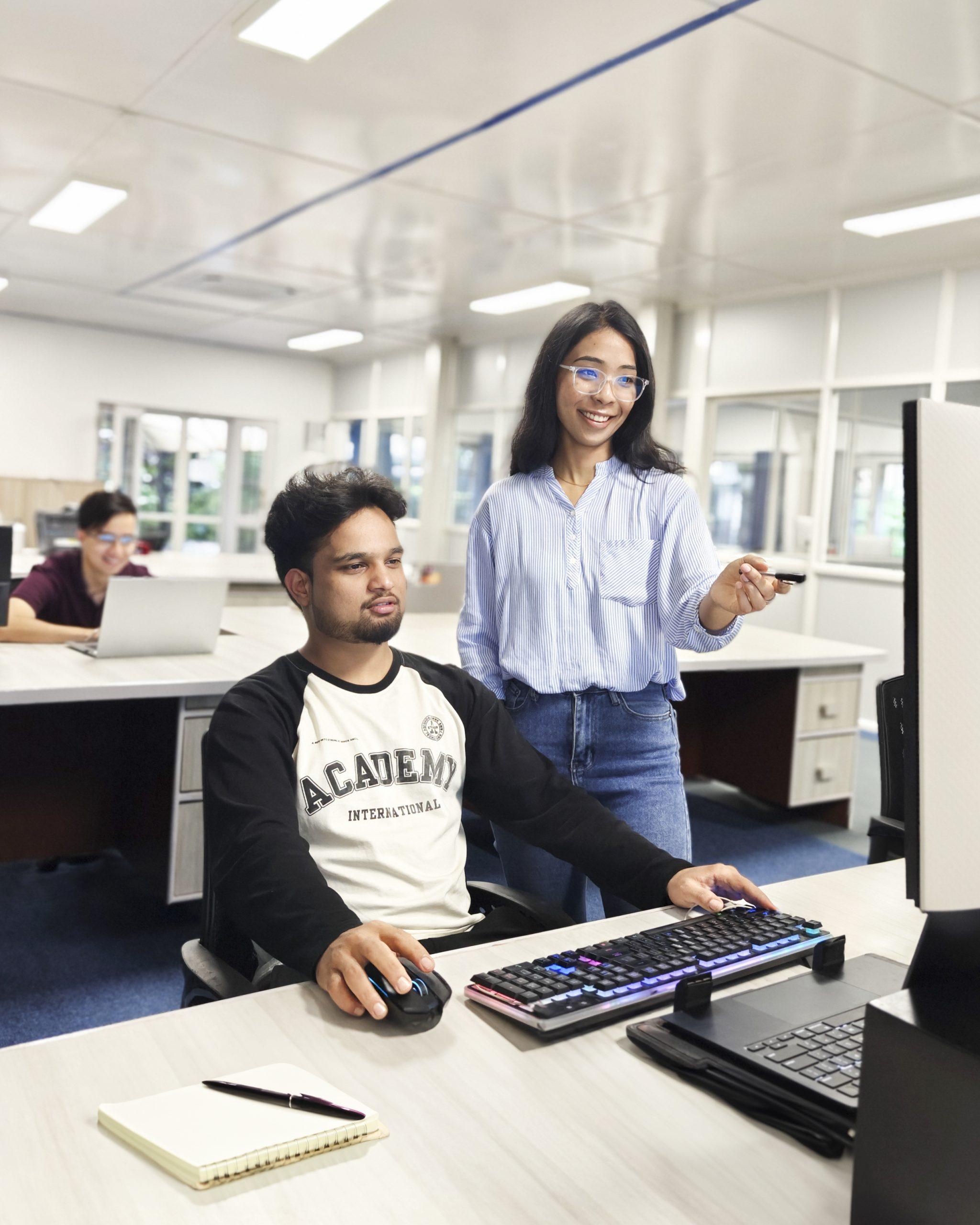 A man sits at a desk using a computer while a woman stands beside him pointing at the screen. They appear to be working together in a bright, modern office with other people in the background.