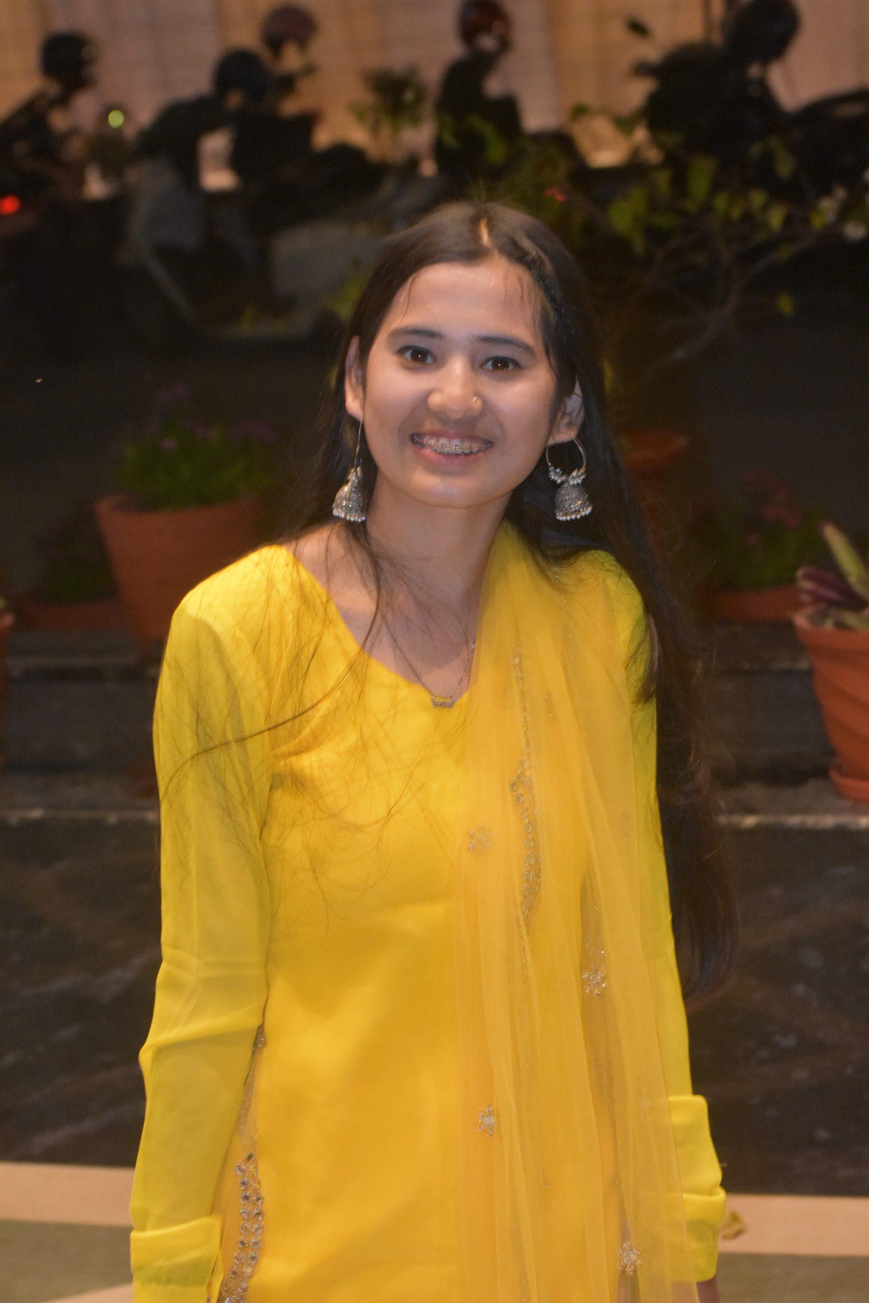 A person wearing a bright yellow traditional outfit and silver earrings is smiling outdoors. They have long hair, and there are potted plants and a dimly lit background behind them.