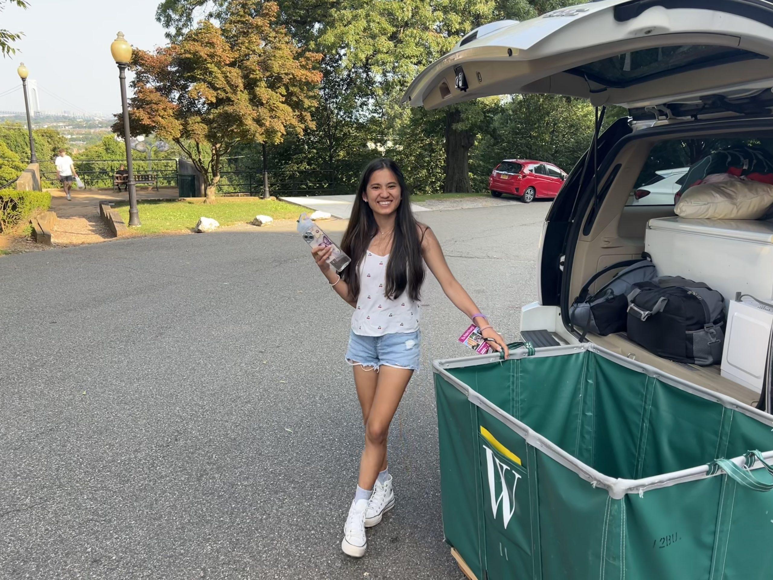 A woman in a sleeveless top and denim shorts stands near an open car trunk filled with bags. She holds a water bottle and snacks, with a large green bin labeled "W" nearby. Trees and parked cars are in the background, on a sunny day.