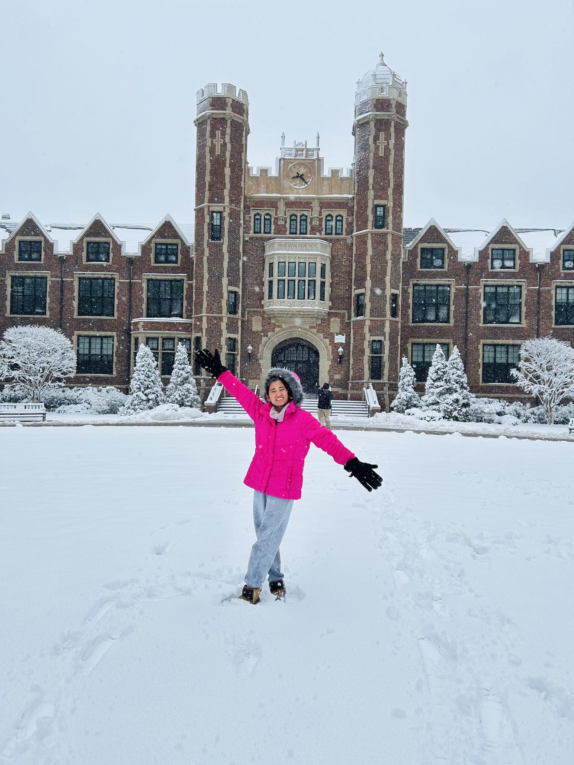 A person in a bright pink coat and gray pants stands on a snowy field, arms raised excitedly. Behind them is a large, historic brick building with snow-covered trees and a clock tower. The scene is wintry and overcast.