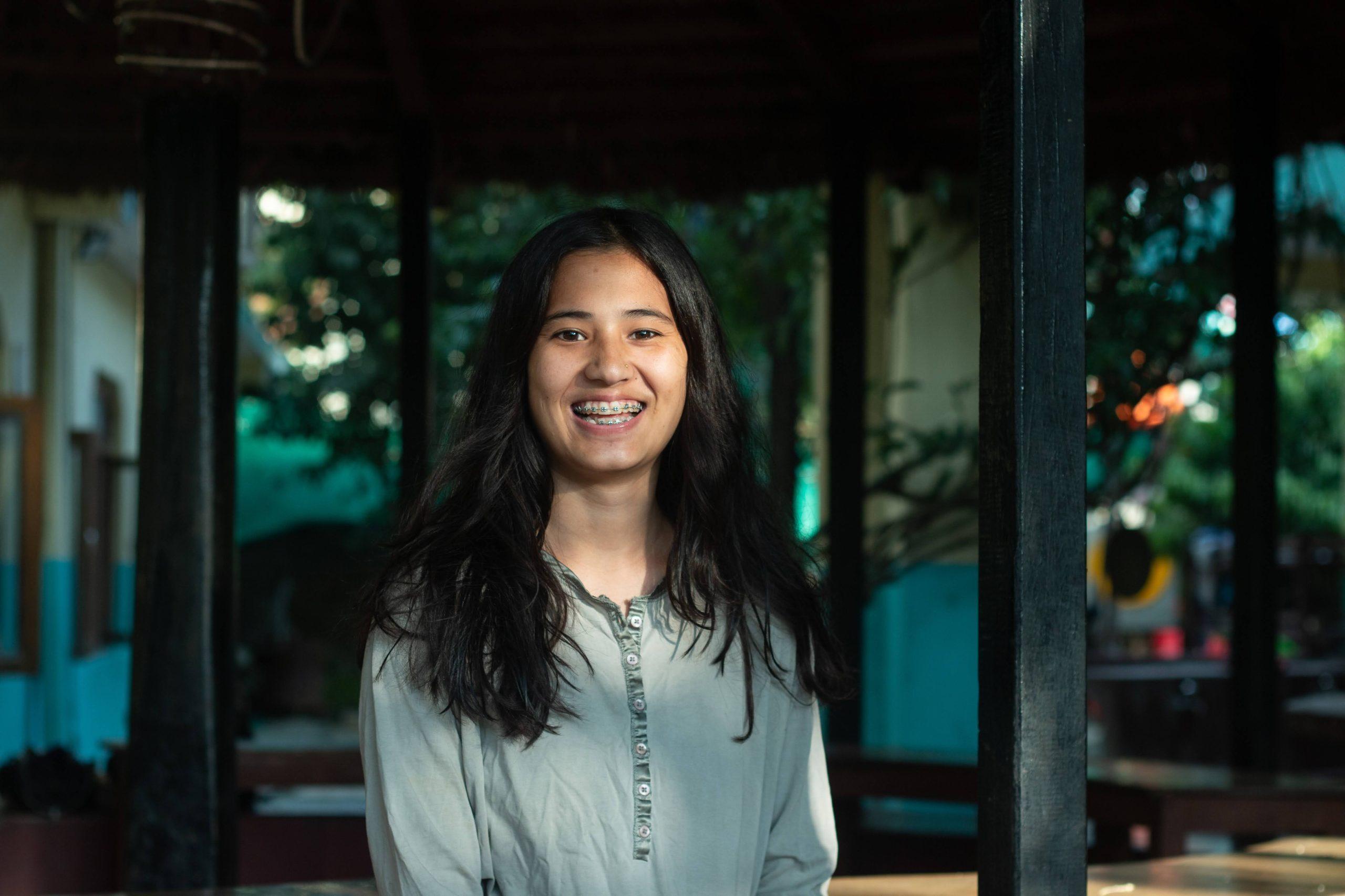 A woman with long, dark hair smiles at the camera. She is wearing a light gray top and standing outdoors under a shaded structure. The background features trees and diffused natural light.