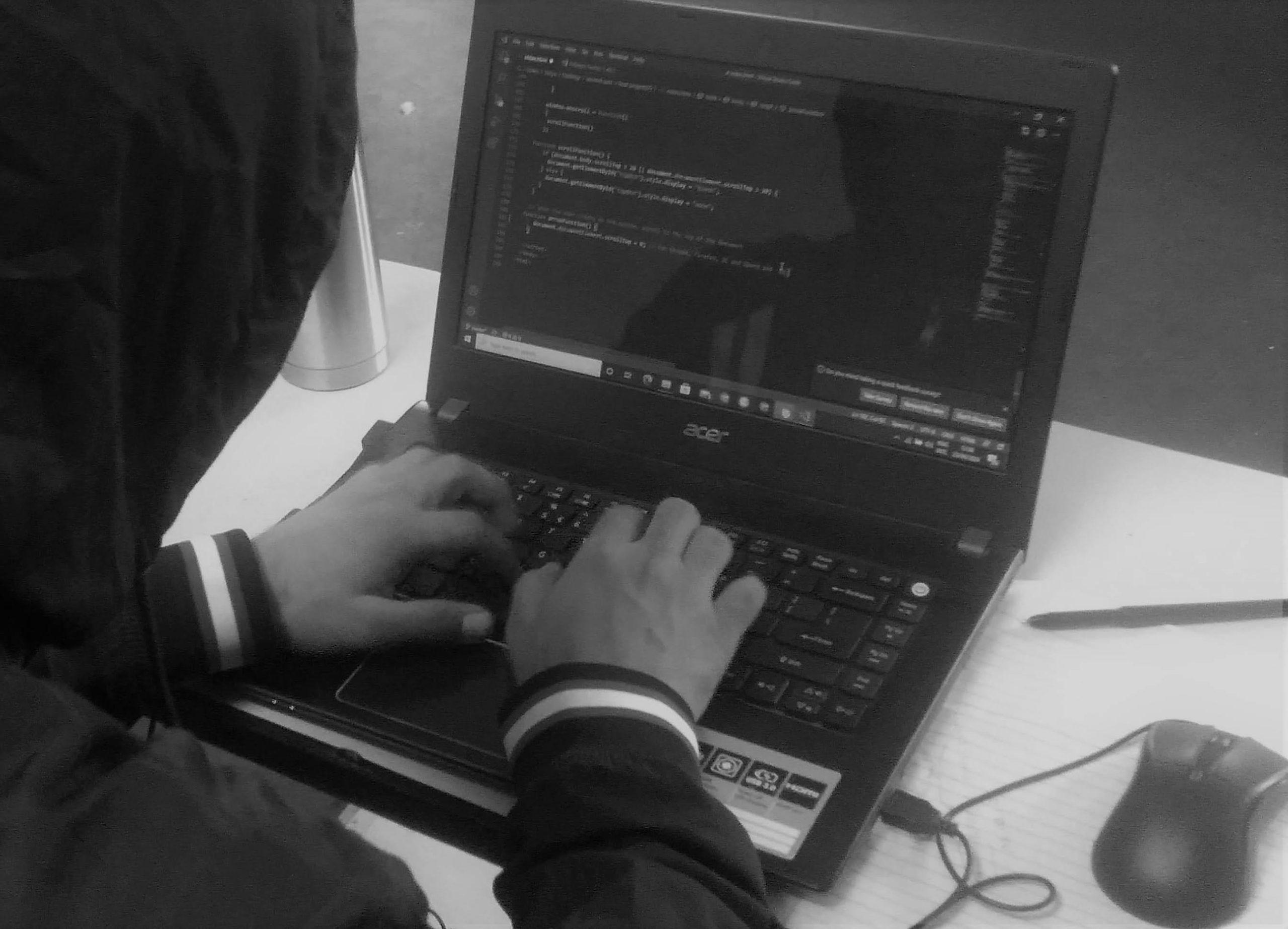 A person in a hooded jacket is typing code on a laptop in black and white. The screen displays lines of code, and a pen and water bottle are on the desk. The setting suggests a focus on programming or computer work.