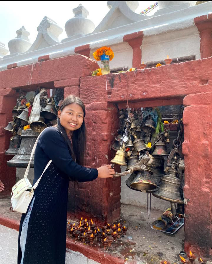 A young woman in a black dress smiles as she rings a large cluster of brass bells at a red stone shrine decorated with flowers and lit oil lamps.