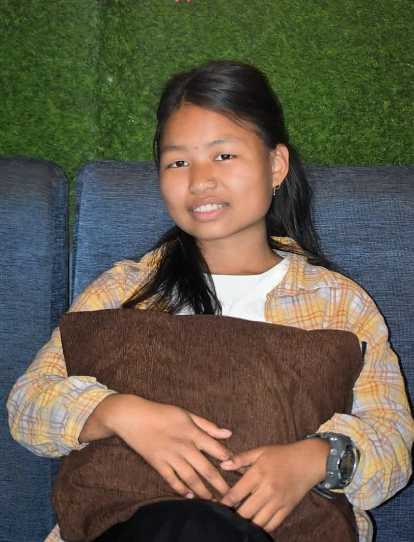 A young woman with long dark hair sits on a blue sofa, holding a brown pillow. She is wearing a yellow plaid shirt over a white top and has a slight smile. The background is green and textured.
