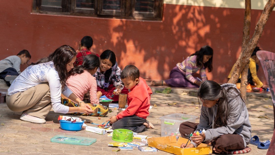 A group of children and a young woman are engaged in a creative activity outdoors. They are seated on a paved area, surrounded by art supplies such as markers and paper. The setting is sunny, casting tree shadows on the ground.