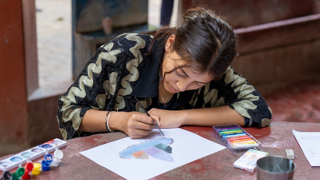 A young girl focuses on painting with watercolors at a wooden table. She wears a patterned jacket and rests her head on her arm as she carefully paints on a sheet of paper. Art supplies are scattered around her.