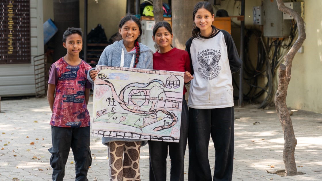 Four children stand outdoors on a paved area, smiling and holding a large drawing of a skate park scene. They are casually dressed, and a tree and building are visible in the background.