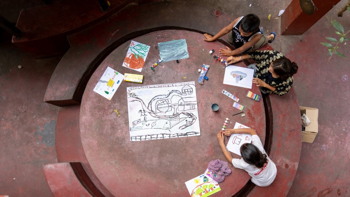 Three children sit around a large, circular table, drawing and painting. The table is cluttered with art supplies, including paints, brushes, and papers. The children appear focused on their artwork in an outdoor setting.