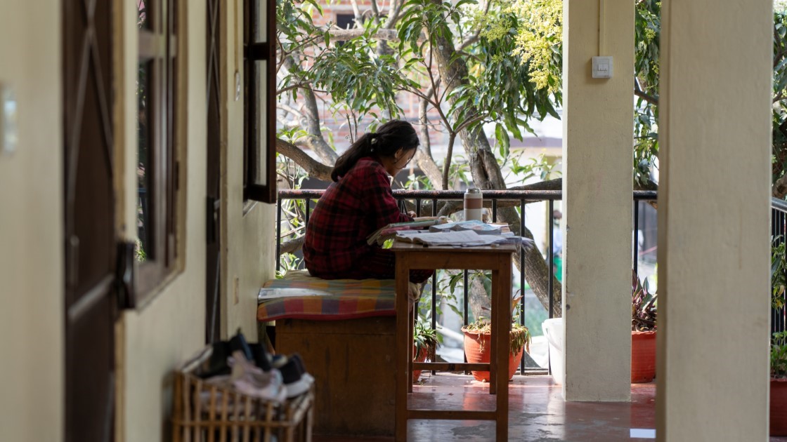 A person sits on a porch, facing away, wearing a red plaid shirt. They are at a wooden table with papers and a drink. Green plants and trees surround the area, and sunlight filters through. A basket of shoes is on the left.